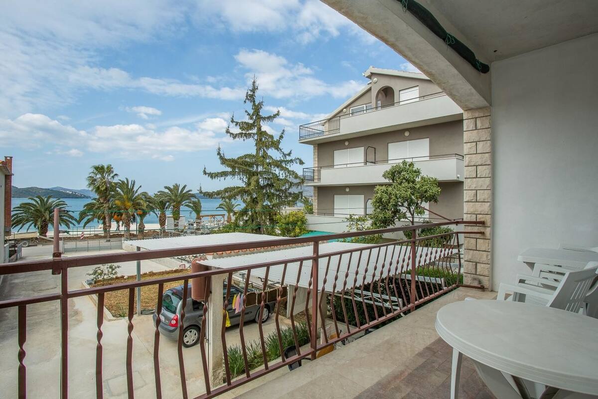 A balcony with a view of the sea, a parked car, and a table with chairs.