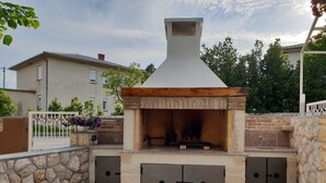 An outdoor pizza oven with a stone wall and metal gates, surrounded by a tiled patio and potted plants. - Apartments Gorana Novi V (Novi Vinodolski)