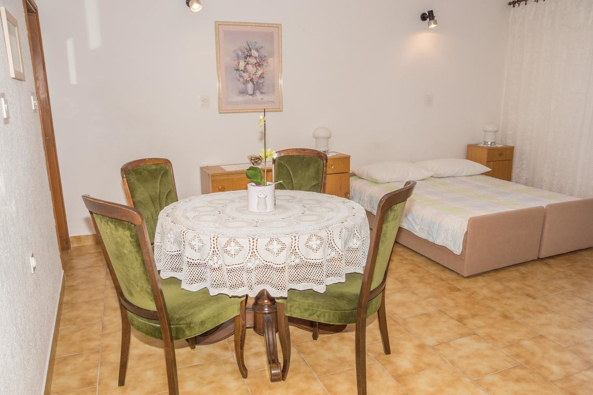 A bedroom with a double bed, a small bedside table, a nightstand, a chair, a table with a lace tablecloth, and a wall-mounted picture.
