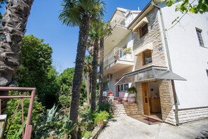 A multi-story building with balconies, a garden with palm trees, and a clear blue sky.