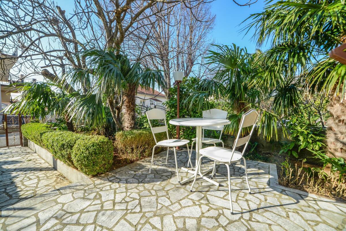 A stone-tiled patio with a round table and two chairs, surrounded by lush greenery and trees.