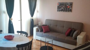 Ein Wohnzimmer mit grauem Sofa, roten und braunen Kissen, einem Glastisch und einer roten Schale auf einer weißen Tischdecke.