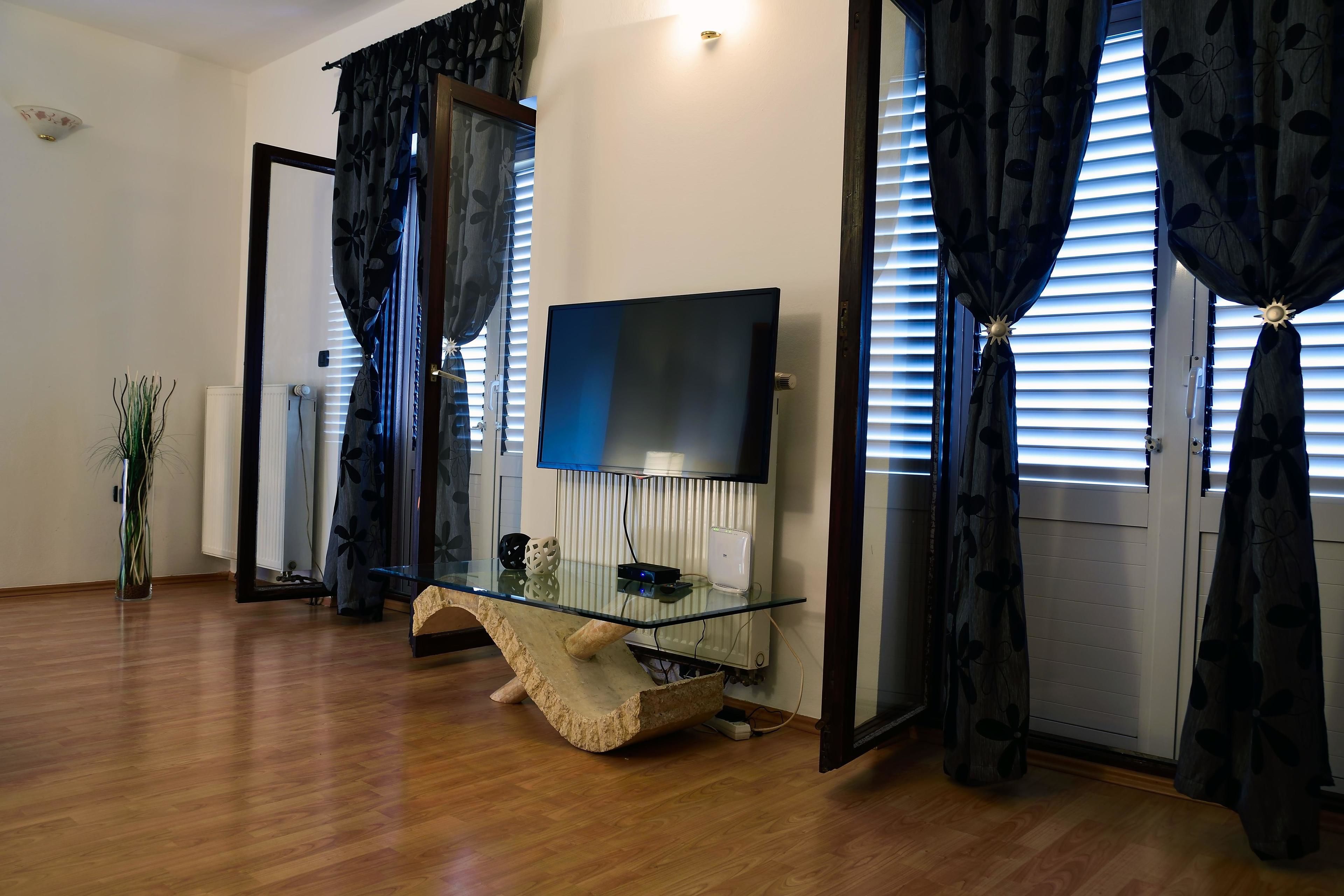 A modern living room with a glass coffee table, a flat-screen TV, and a large window with blinds.