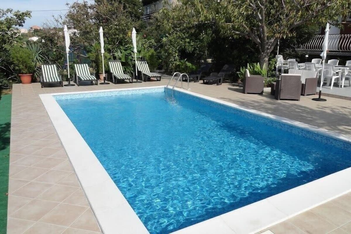 A rectangular swimming pool with clear blue water, surrounded by a tiled deck. There are several lounge chairs and umbrellas, and a small garden area with potted plants.