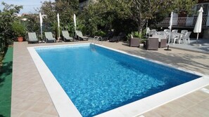 A rectangular swimming pool with clear blue water, surrounded by tiled flooring and outdoor furniture including lounge chairs and umbrellas.
