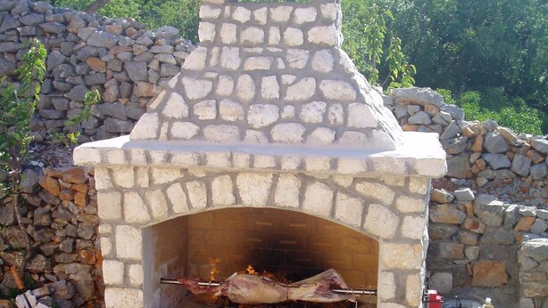 A stone-built outdoor oven with a chimney, surrounded by a stone wall and greenery. - Apartments Biondić (Senj)