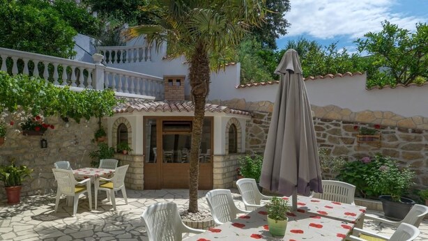 A patio area with a table set for two, surrounded by white chairs, a large umbrella, and potted plants. - Apartments Pribil (Rab)