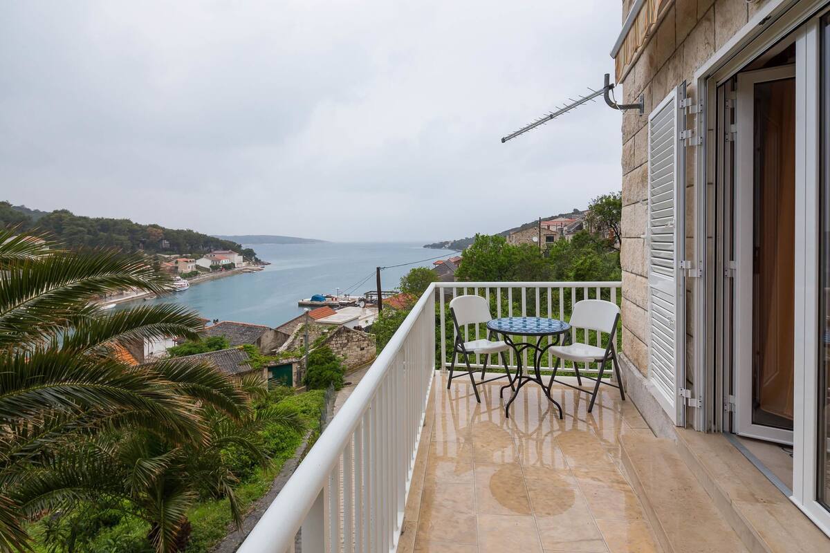A balcony with a table and chairs, overlooking a coastal view with buildings and a boat.