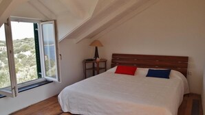 Un dormitorio con piso de madera, una cama con cubrecama blanco, almohada roja y azul, mesita de noche con lámpara y ventana con vista a follaje.