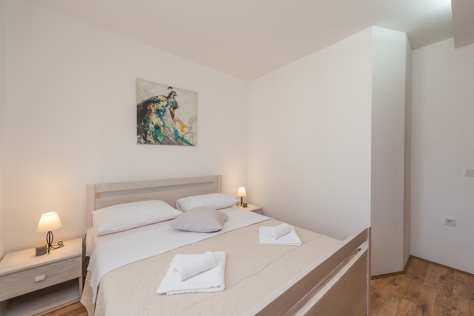 A neatly made bed with white linens, two bedside lamps, a wall-mounted abstract painting, and a wooden floor.