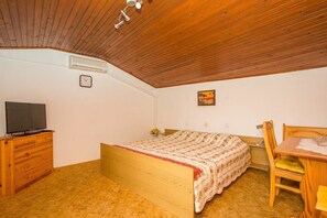 A bedroom with a wooden ceiling, a bed with a floral bedspread, a wooden dresser with a television, a small table with a chair, and a wall clock.