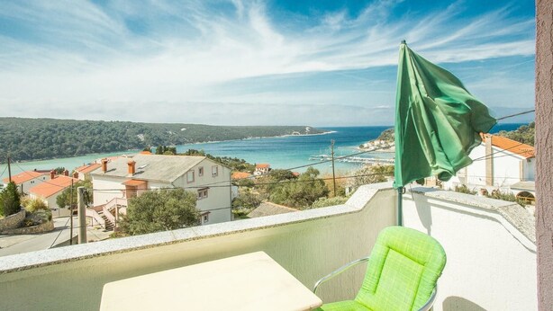 A balcony with a view of a coastal town, a green umbrella, and a white table. - Apartments Antonija (Rab)
