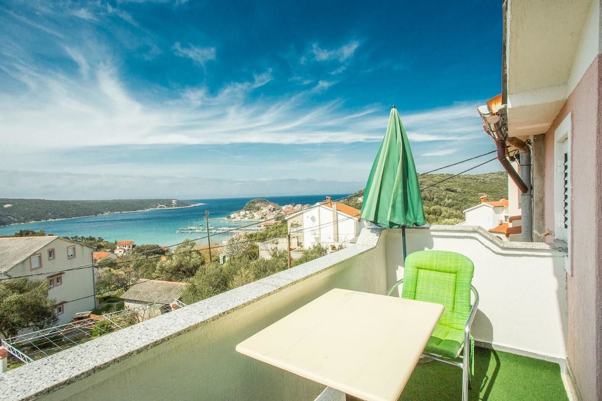 Un balcón con una mesa y una silla, con vistas a un pueblo costero bajo cielos despejados.