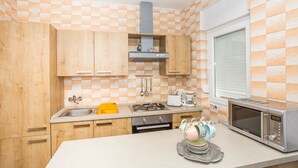 A kitchen with wooden cabinets, a sink, a stove, a microwave, and a window with blinds. - Apartments Antonija (Rab)