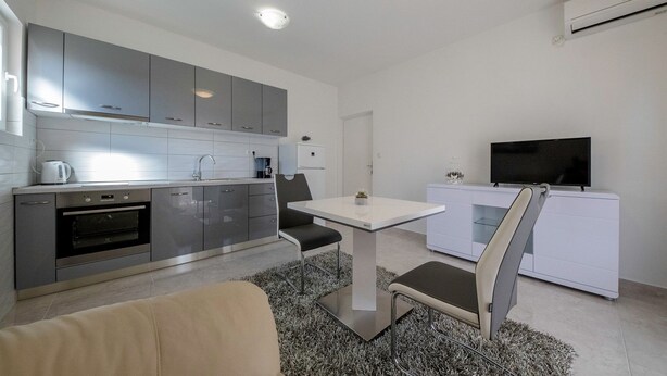 A modern kitchen with grey cabinets, a built-in oven, and a dining area with a white table and two chairs. - Apartments Marica 3131 (Rab)