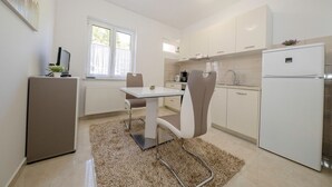 A modern kitchen with white cabinets, a refrigerator, a dining table with chairs, and a window with curtains. - Apartments Marica 3131 (Rab)