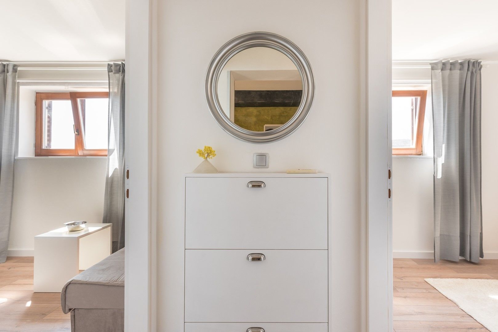 A modern bedroom with a white dresser, a round mirror, and a small vase with flowers.