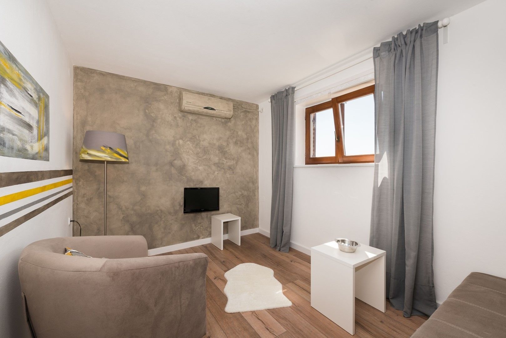 A modern living room with a flat-screen TV, a small white side table, a beige sofa, a white rug, and a window with grey curtains.