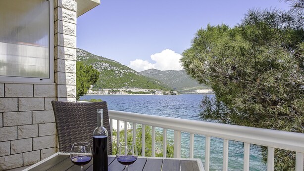 A balcony with a table, two wine glasses, and a bottle of wine. The view includes a body of water and green hills in the background. - Apartments Krstičević (Slivno)