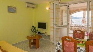 A room with a sofa, a TV, a small table with a vase, and a dining area with chairs and a table set with fruit. - Apartments Krstičević (Slivno)