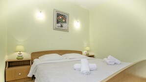 A neatly made bed with white linens and rolled towels, two bedside lamps, a nightstand, and a framed painting on the wall. - Apartments Krstičević (Slivno)