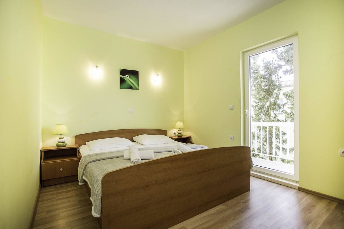 A bedroom with a wooden bed, two bedside tables with lamps, a small wall-mounted picture, and a large window with a view of trees.