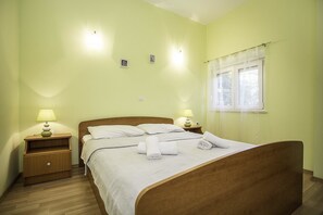 A neatly made bed with white linens and rolled towels, a wooden nightstand with a lamp, and a window with sheer curtains. - Apartments Krstičević (Slivno)