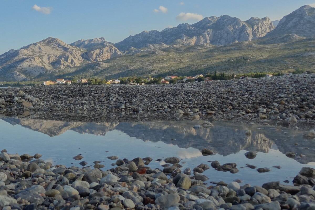 A rocky shoreline with a calm body of water reflecting the surrounding landscape, including distant mountains and a few buildings.