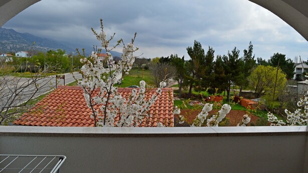 A view through an arched window of a building with a red-tiled roof, a garden with blooming trees, and a mountainous landscape in the background. - Apartments Ante Seline (Starigrad)