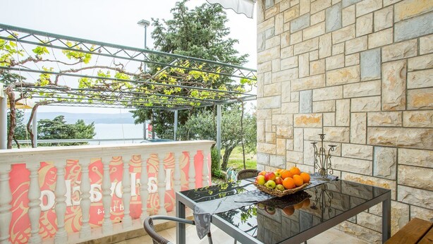 A balcony with a stone wall, a glass table with fruit, and a metal railing. - Apartments Divna Tomas (Omis)