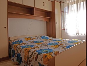 A bedroom with a bed, wooden headboard, built-in shelving, and a window with curtains. - Apartments Janja Postira (Postira)