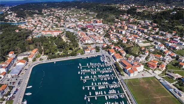An aerial view of a coastal town with a marina, residential buildings, and a large building with a red roof. - Holiday House Mali raj (Rab)