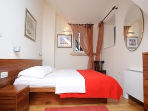 A bedroom with a wooden bed, a red blanket, a bedside table, a mirror, and a window with curtains. - Apartments Vila Marija (Supetar)