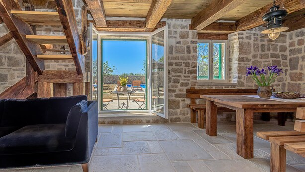 Un interior rústico con pared de piedra, vigas de madera, un sofá, una mesa y vistas a una piscina.