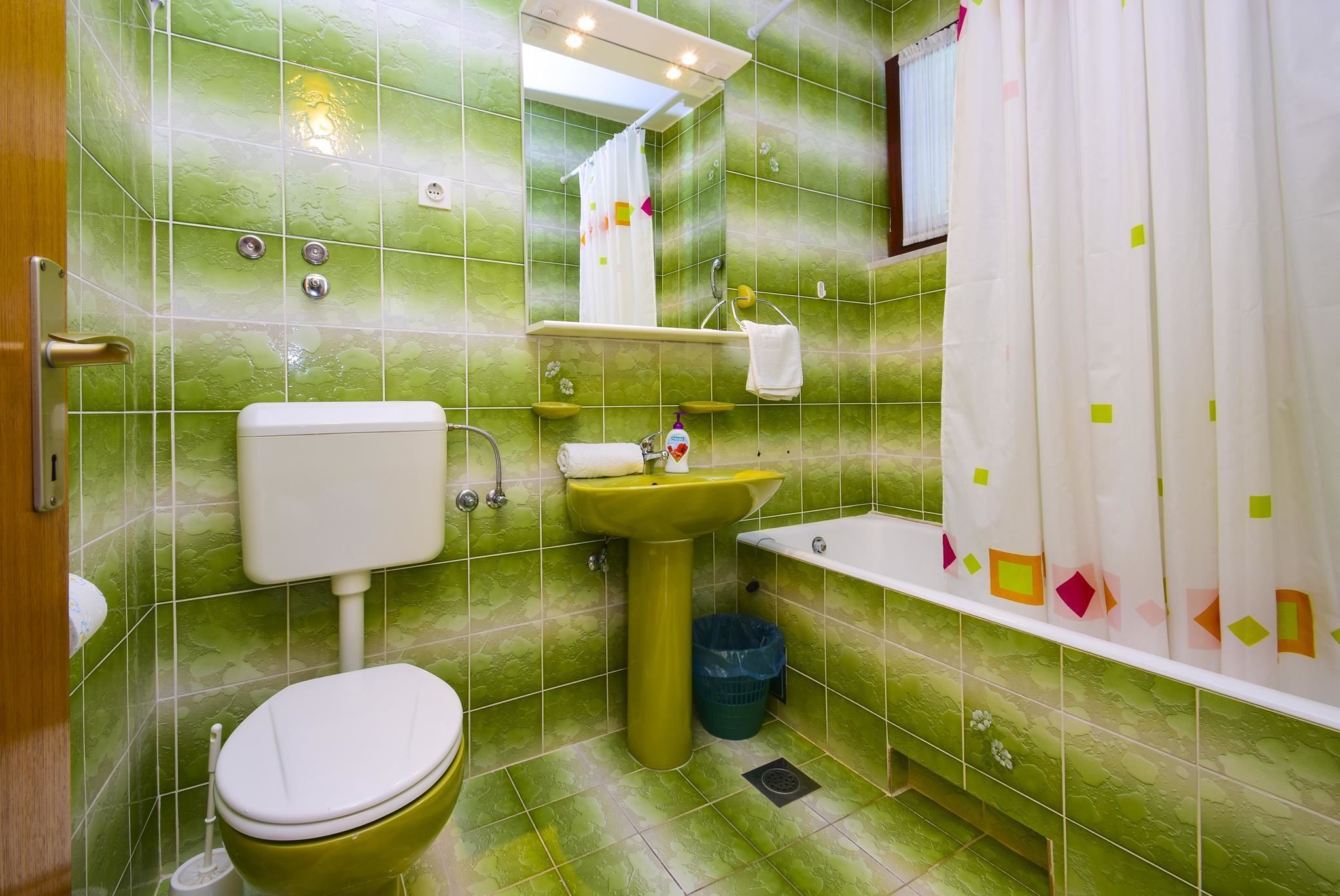 Et badeværelse med grønne fliser, toilet, håndvask og badekar.