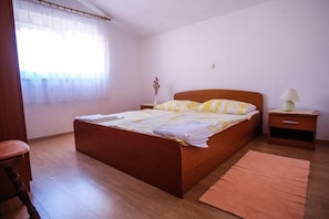 Ein Schlafzimmer mit einem hölzernen Bett, einem Nachttisch mit Lampe, einem Fenster mit durchsichtigen Vorhängen und einem Holzboden.