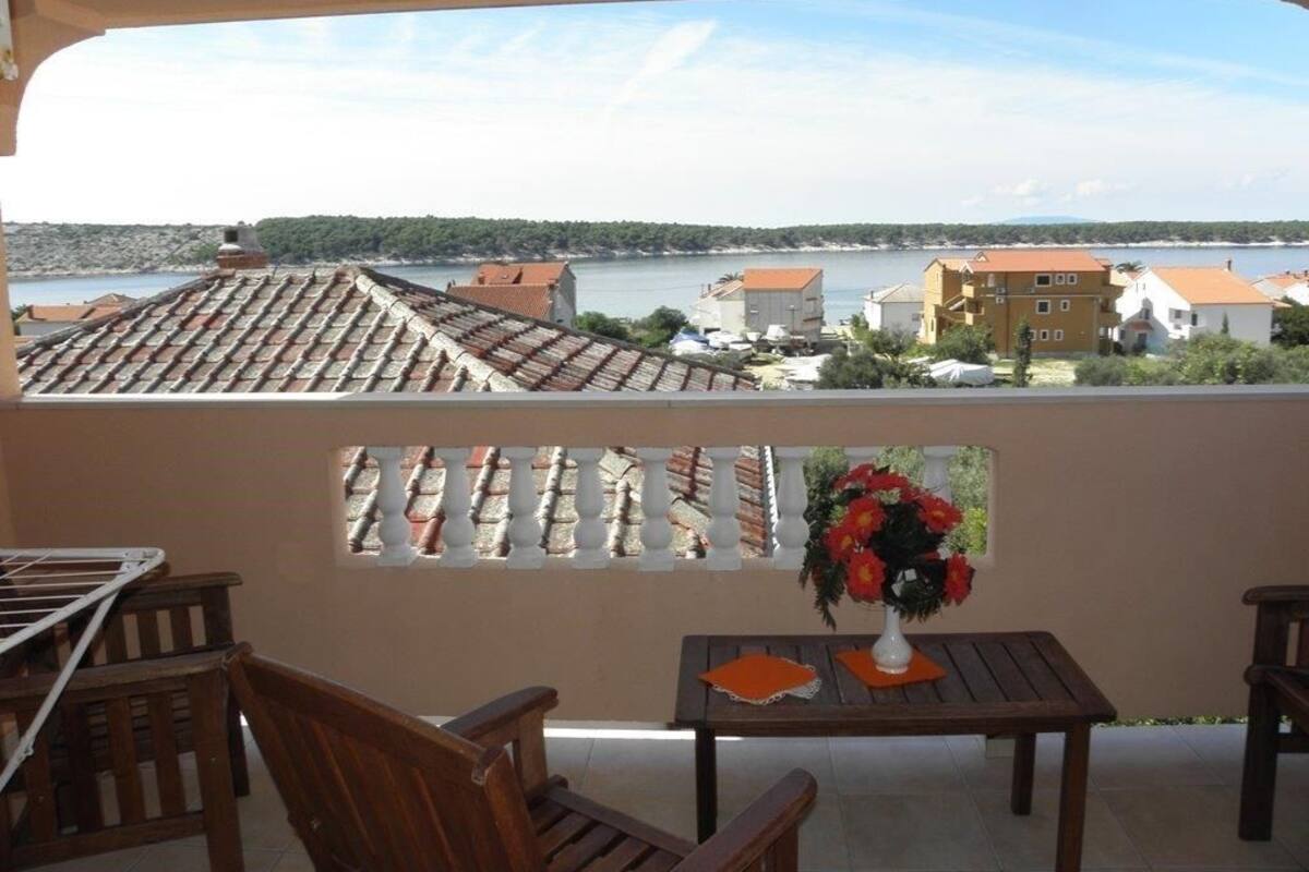 Un balcon donnant sur la côte, avec une table ornée d’un vase de fleurs, des chaises en bois et une balustrade.