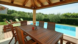 Eine Holzterrasse im Freien mit Billardtisch und Stühlen, umgeben von einem Pool und viel Grün.