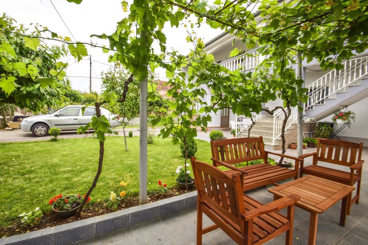 A patio with wooden furniture, a small garden, and a building with a staircase in the background.