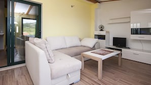 Ein Wohnzimmer mit einem weißen Sofa, einem Holztisch, einem Fernseher auf einem Schrank und einer Glasschiebetür, die auf einen Balkon führt.