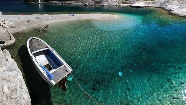 Un bateau amarré à une côte rocheuse, avec une eau turquoise et limpide, une plage de sable et une colline boisée en arrière-plan.