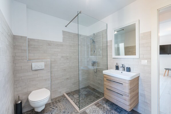 A modern bathroom with a glass-enclosed shower, a white toilet, a wooden vanity with a mirror, and patterned floor tiles.