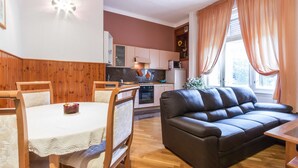 A cozy living room with a brown leather sofa, wooden dining table, and chairs. A kitchen with white cabinets and a microwave is visible in the background.