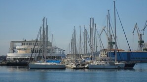 A marina with several sailboats docked, a building with a modern design, and cranes in the background.