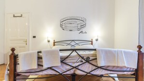 A bed with a metal frame, white bedding, and two towels. A wall decoration of a coliseum is visible above the bed.