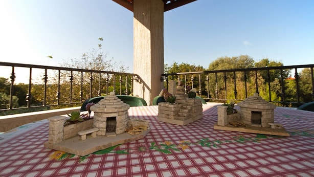 A balcony with a checkered tablecloth, two stone structures resembling fireplaces, and a view of trees and a fence. - Apartment Gasparini / Three Bedrooms (Pula)