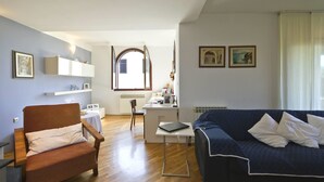 A living room with a wooden floor, a blue sofa, a brown armchair, a white desk, and a window with a view of a building. - Apartment Gasparini / Three Bedrooms (Pula)