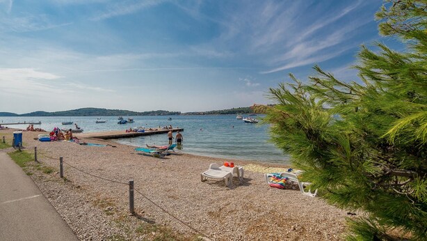 A pebbled beach with lounge chairs and a dock extending into calm water. - Apartments Grulović (Vodice)