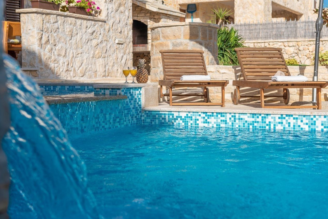 Un espace piscine comprenant deux chaises longues en bois, un mur en pierre et une bordure carrelée.