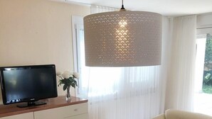 A living room with a television on a wooden cabinet, a beige sofa, a round white coffee table with a vase of flowers, and a large pendant light. - Holiday House Lidija (Medulin)